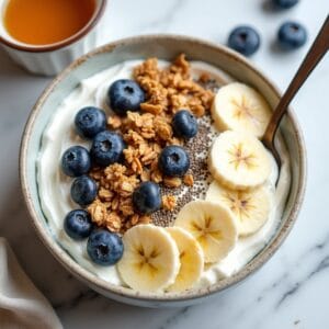 Closeup de uma tigela de iogurte natural ou açaí, coberta com uma explosão de texturas e cores: granola caseira, fatias de banana, mirtilos, sementes de chia e um fio de mel. A imagem deve ser vibrante e apetitosa, focando na riqueza dos ingredientes que promovem a saúde intestinal e o bemestar.
